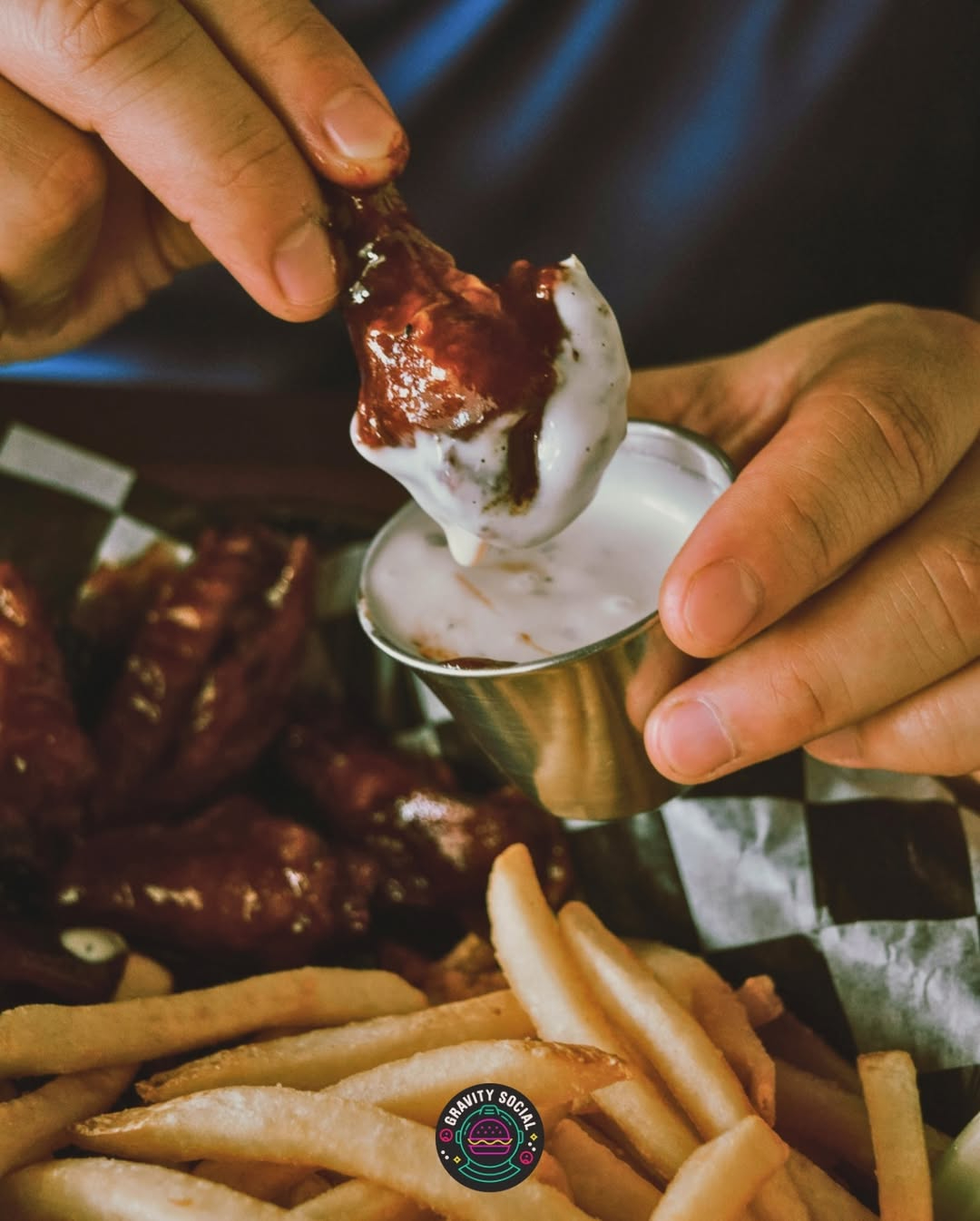 Close-up of a hand dipping a saucy chicken wing into a cup of ranch with fries on the side.