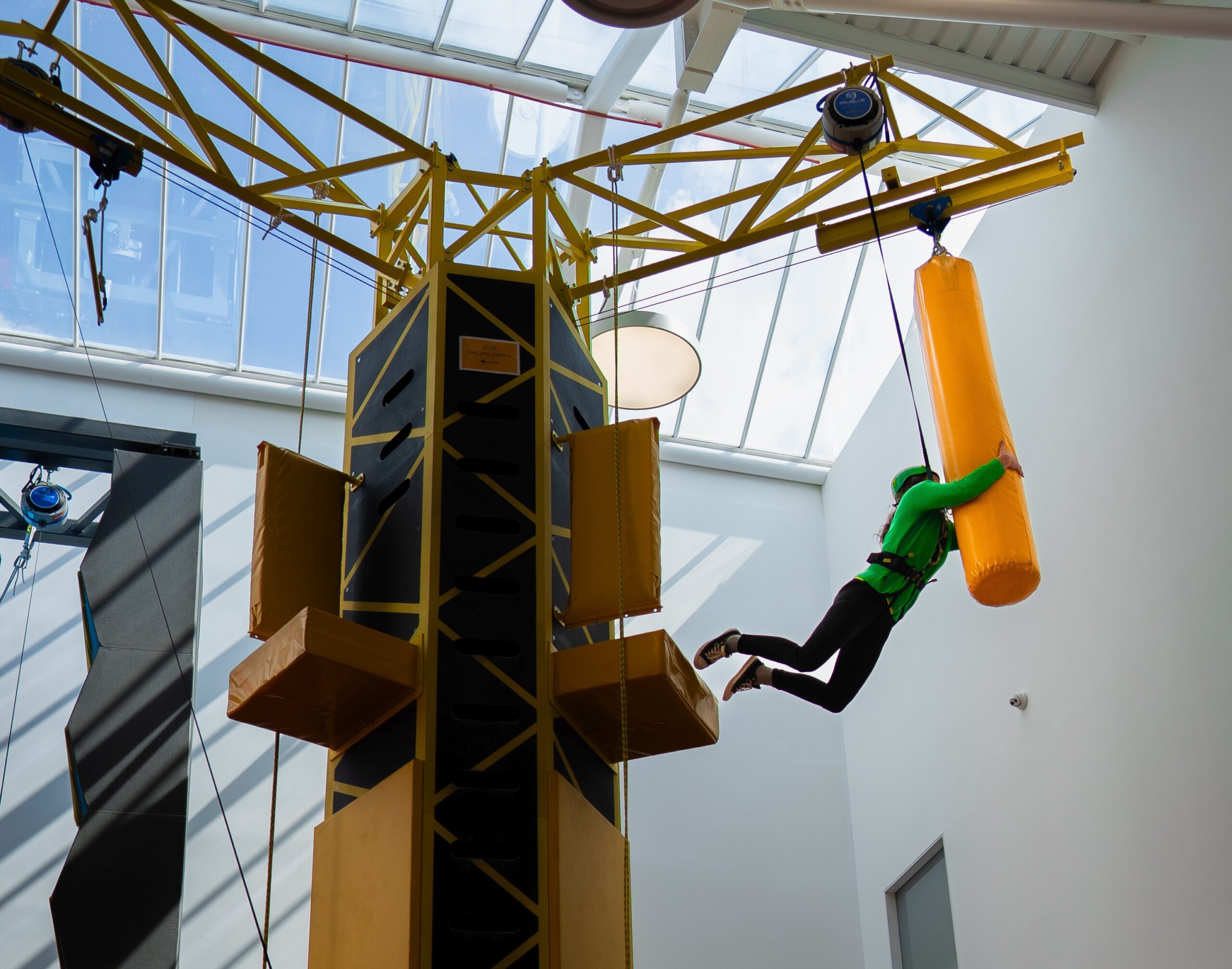 Person in harness climbing indoor adventure course with yellow obstacle, bright lighting.
