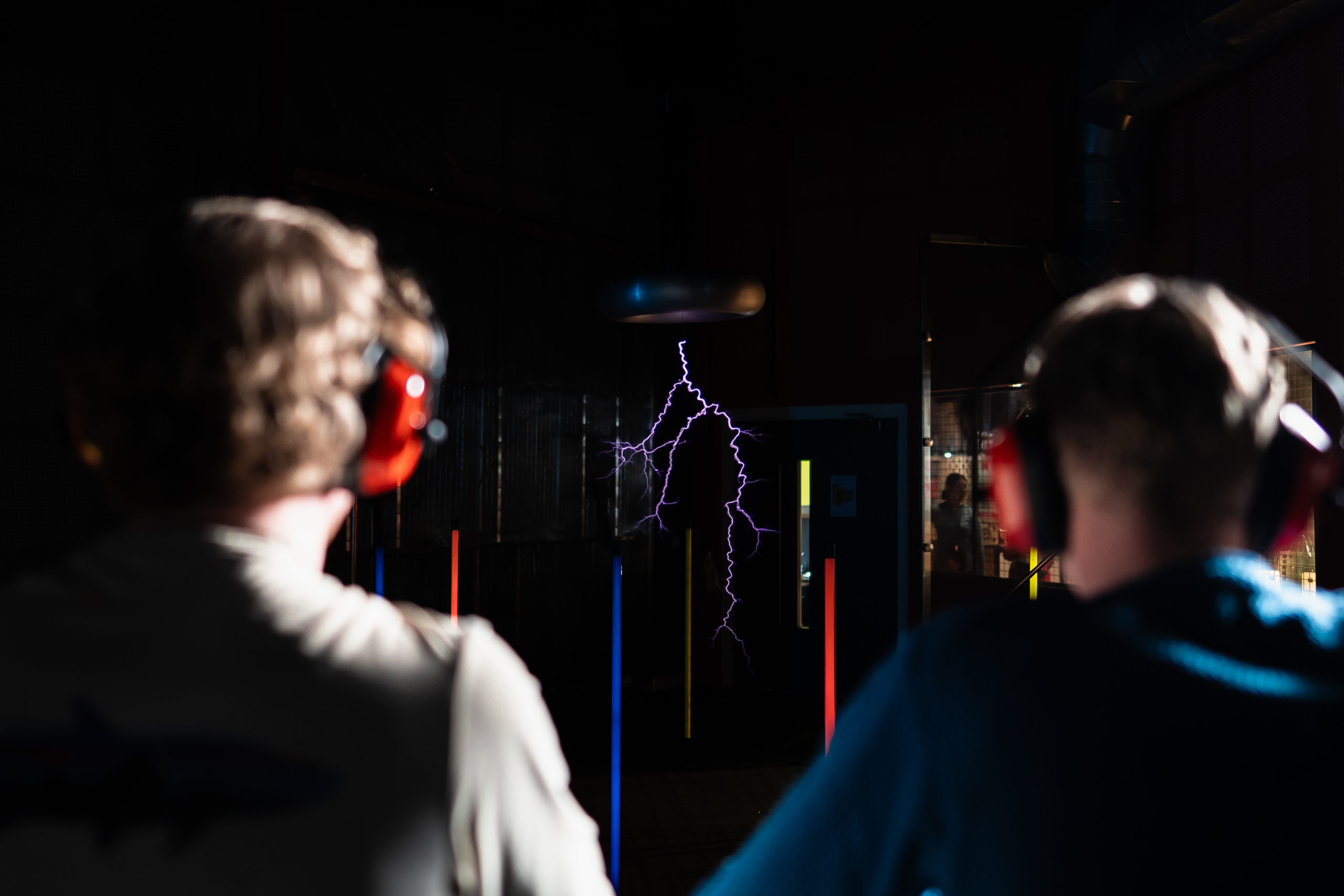 Two people wearing ear protection watch a dramatic indoor lightning exhibit in a dimly lit room.