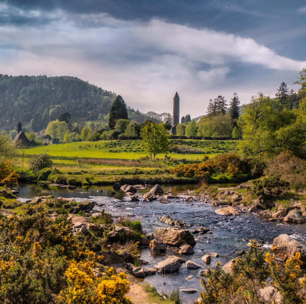 Medieval tower in scenic Irish landscape with mountains, river, and lush greenery under a cloudy sky.