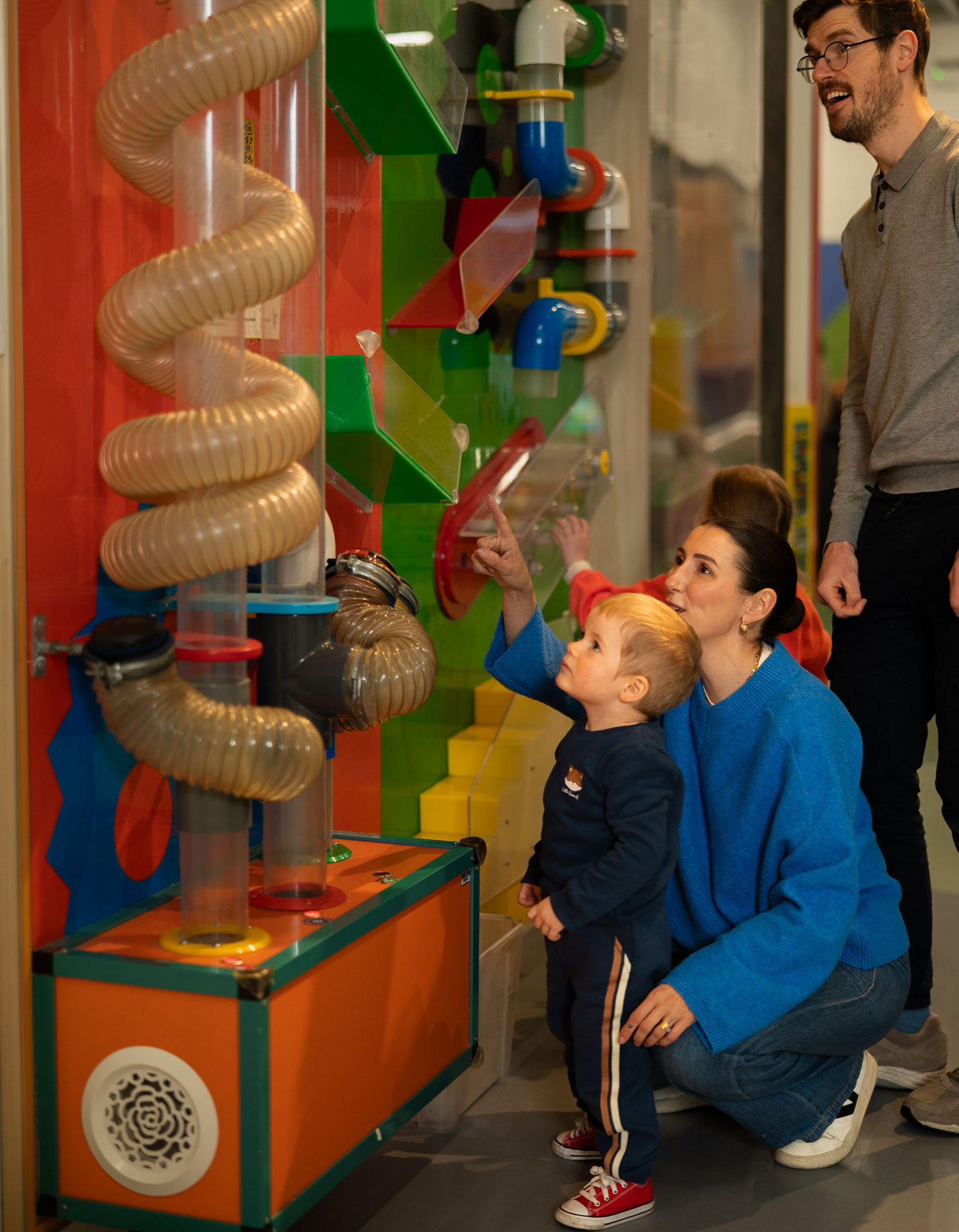 Child exploring an interactive exhibit with family at a science museum. Colorful tubes and gears in background.