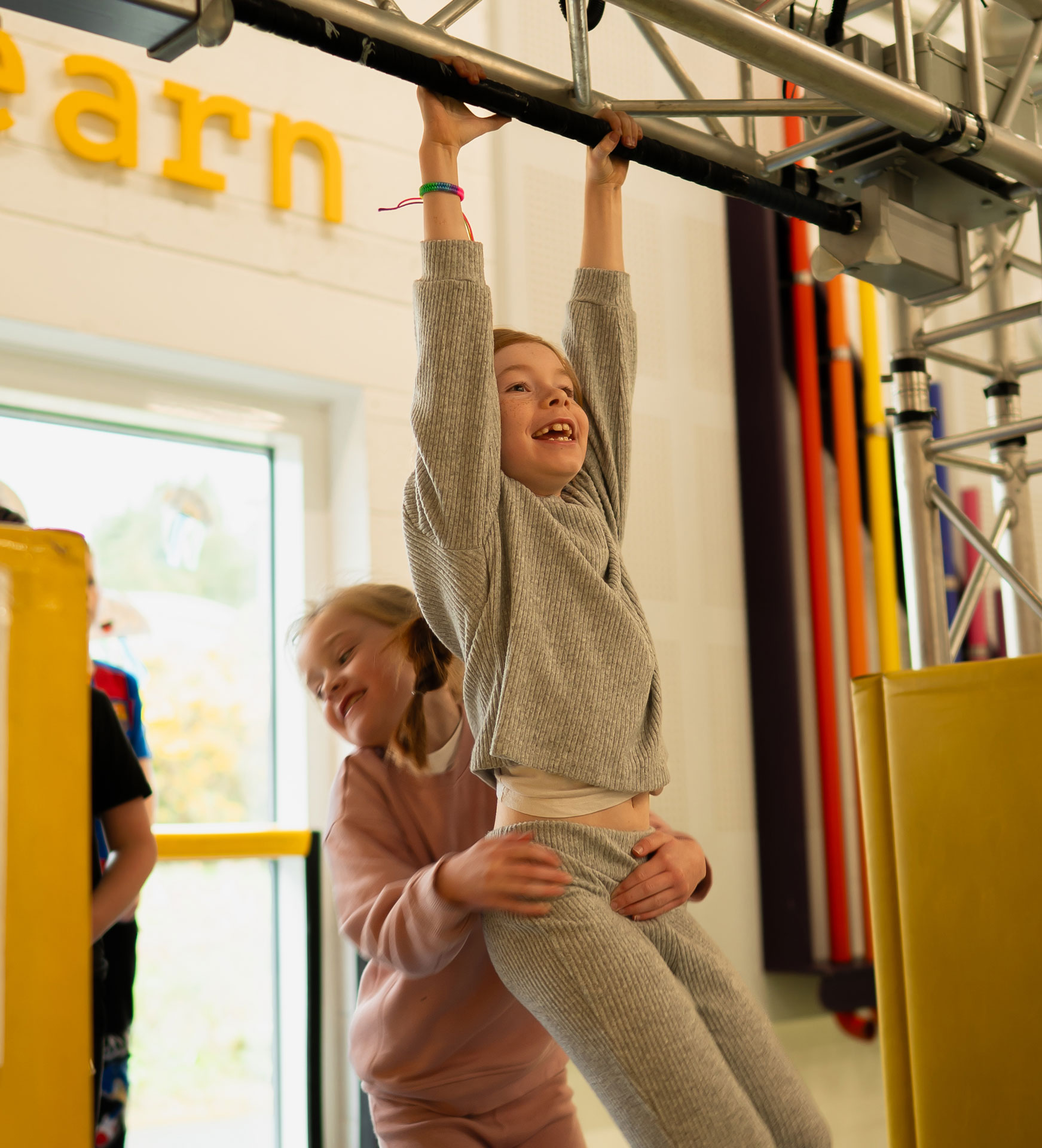 Children playing on indoor monkey bars at a learning center, smiling and helping each other.