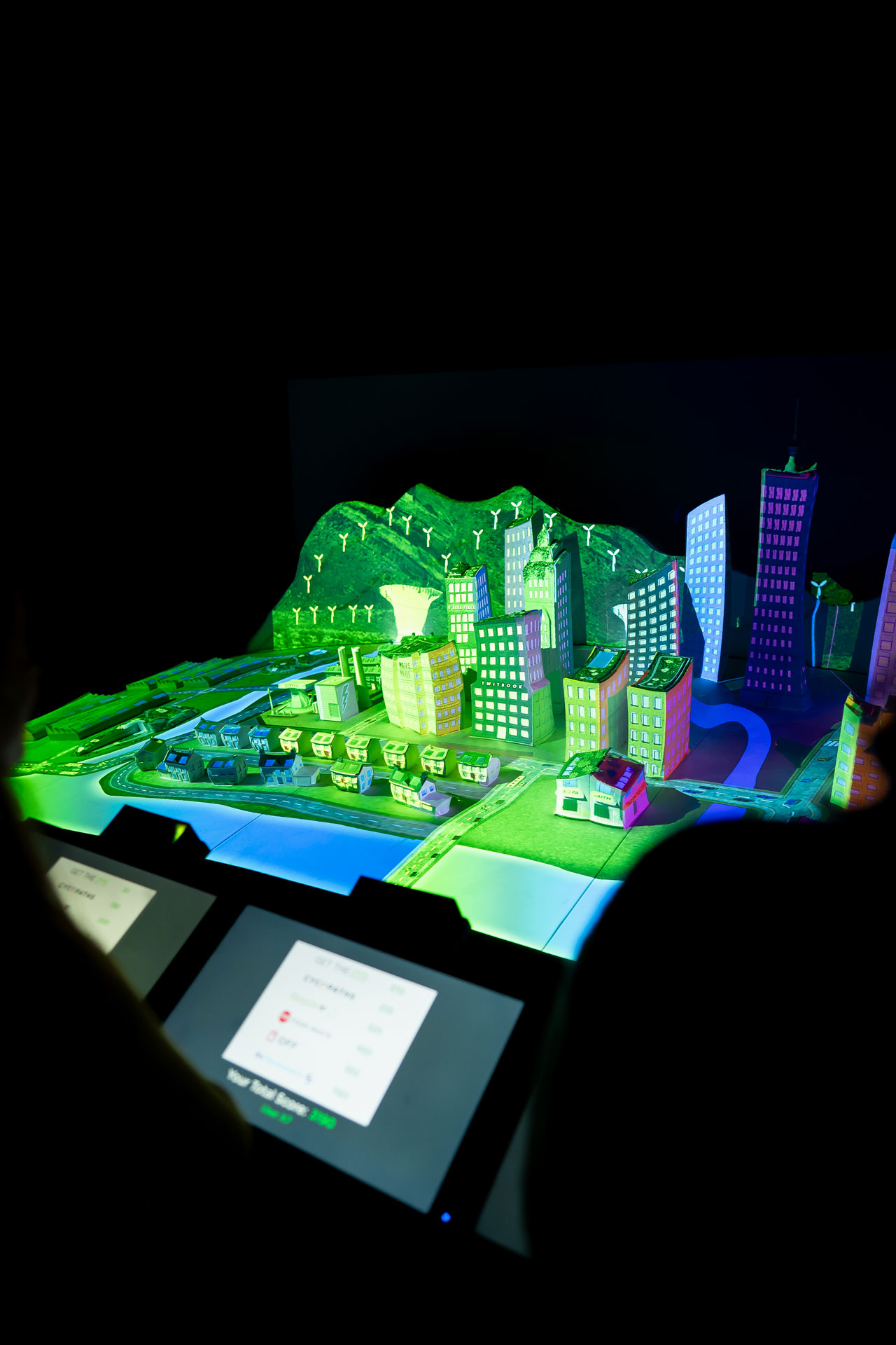Illuminated futuristic city model with wind turbines and vibrant buildings on display panel.
